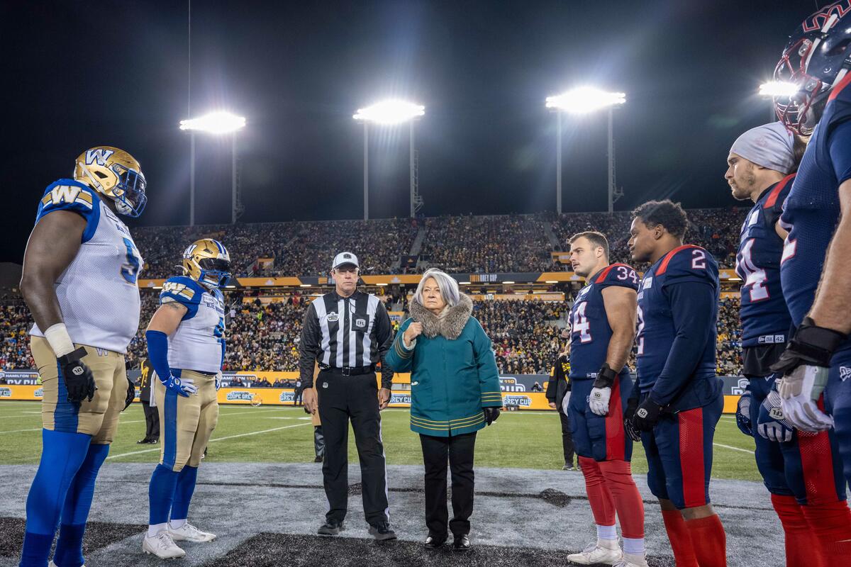 Governor General Mary Simon tosses a coin a surrounded by football players and the referee