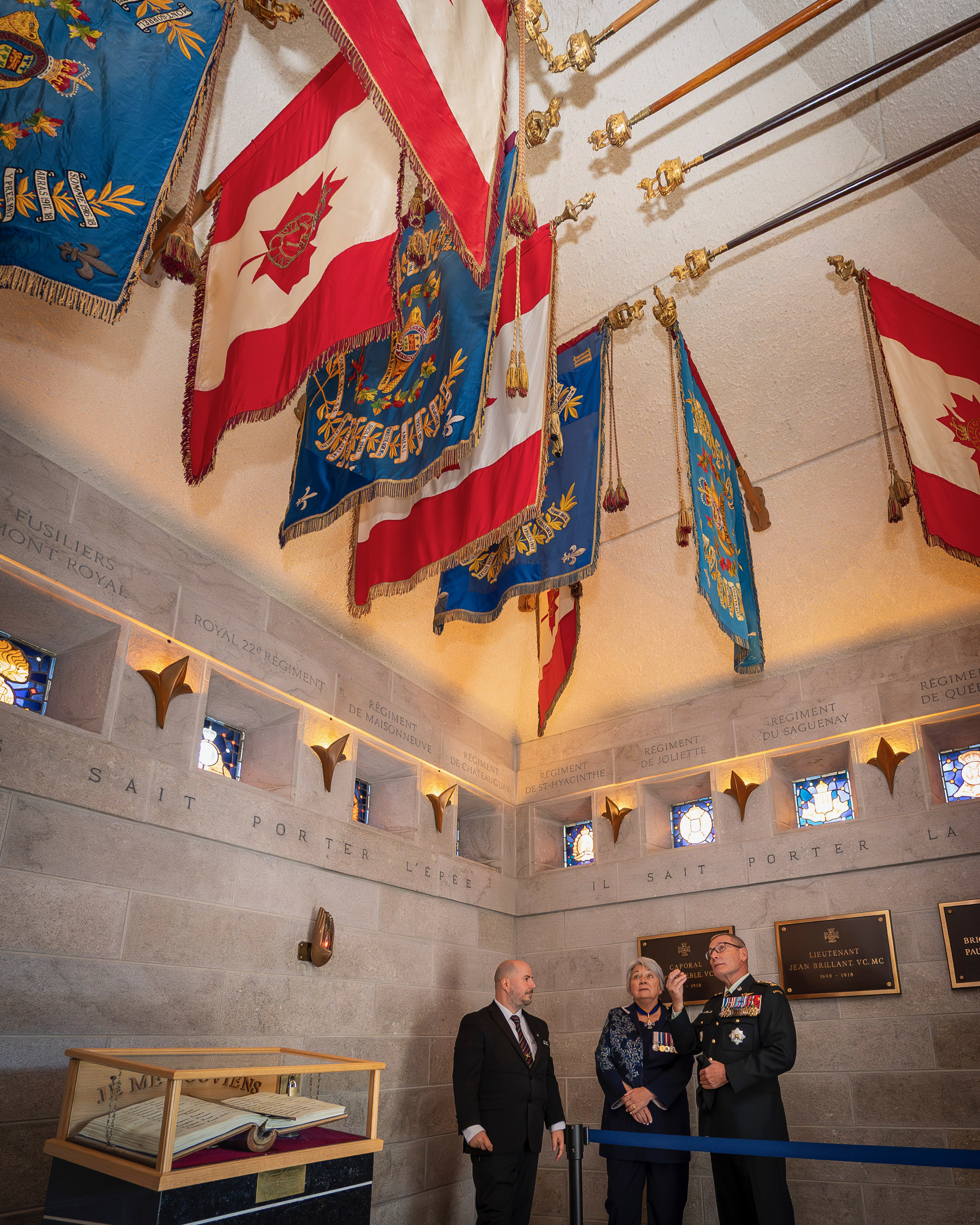 La gouverneure générale Mary Simon debout avec deux personnes sous des drapeaux colorés et ornés suspendus au plafond dans une salle solennelle. Des plaques ornent les murs.