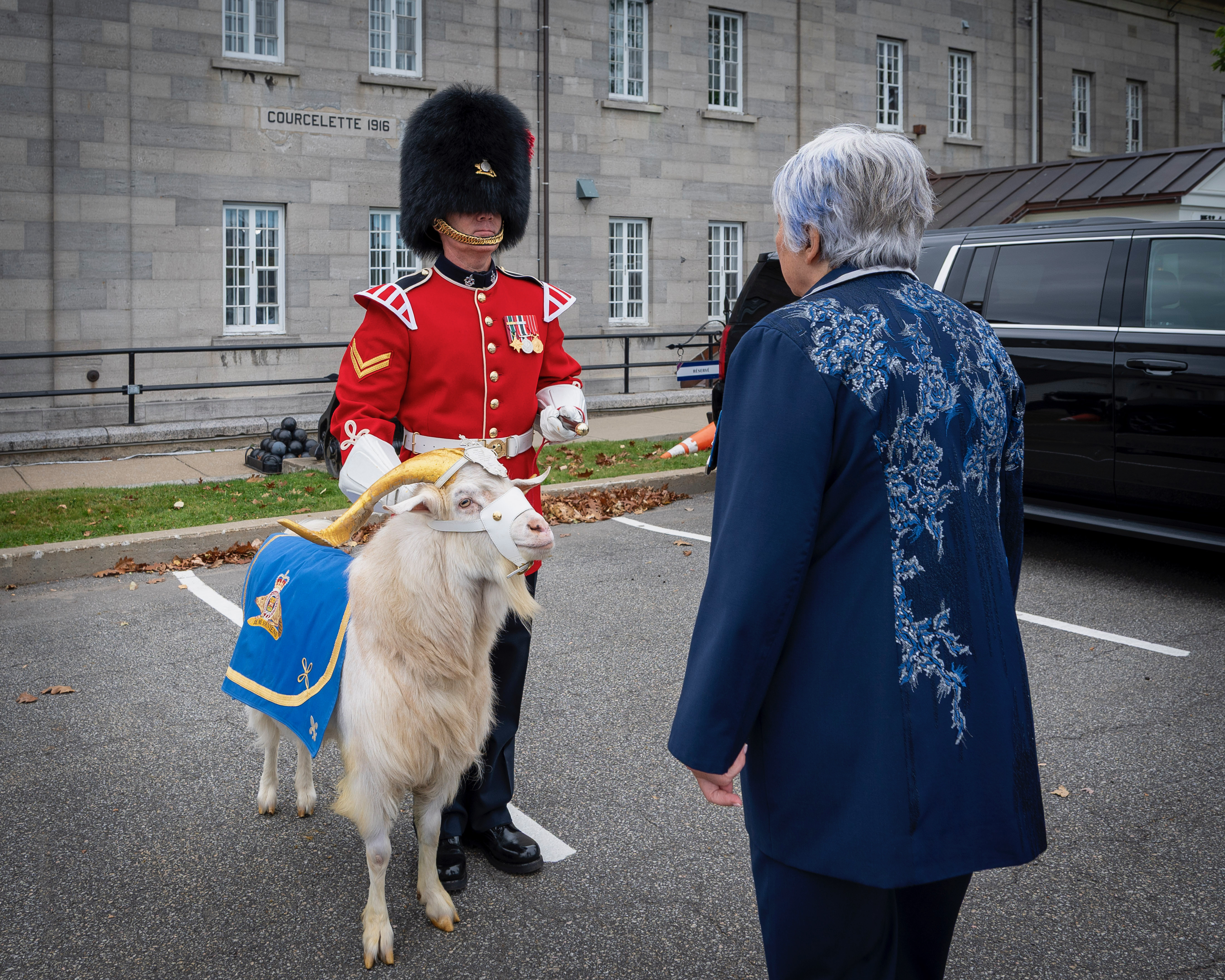 La gouverneure générale Mary Simon debout devant un soldat en uniforme cérémoniel rouge, coiffé d'un haut chapeau noir et tenant une chèvre blanche drapée d'un tissu bleu.