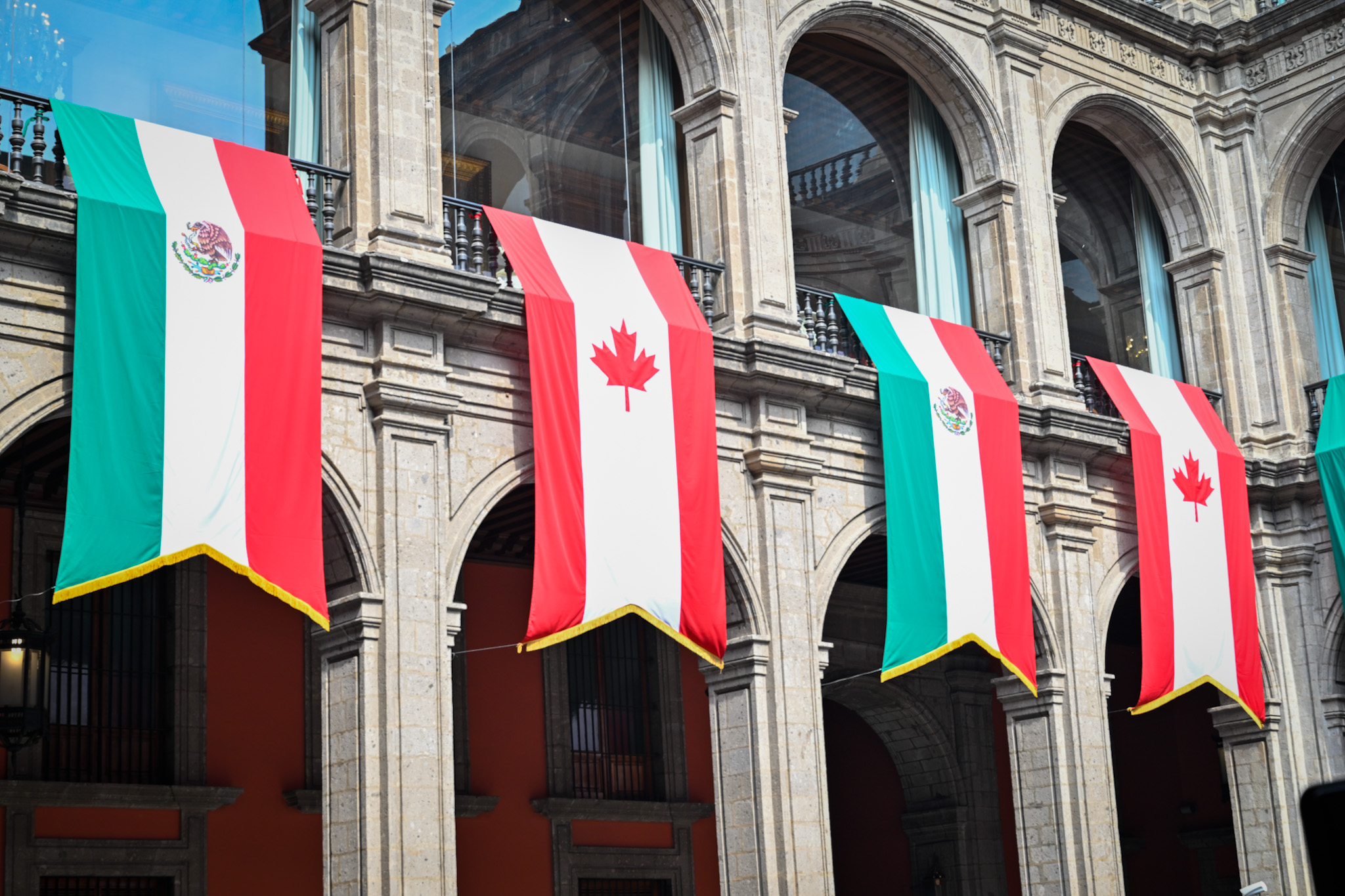Les drapeaux du Mexique et du Canada sont accrochés en alternance sur un bâtiment historique en pierre aux fenêtres cintrées.