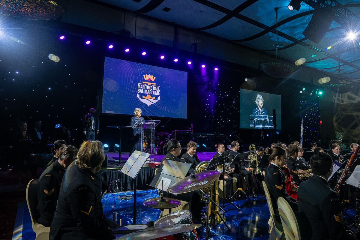 Governor General Arbour standing at the podium a the Maritime Ball while a band with naval attire and instruments listen.