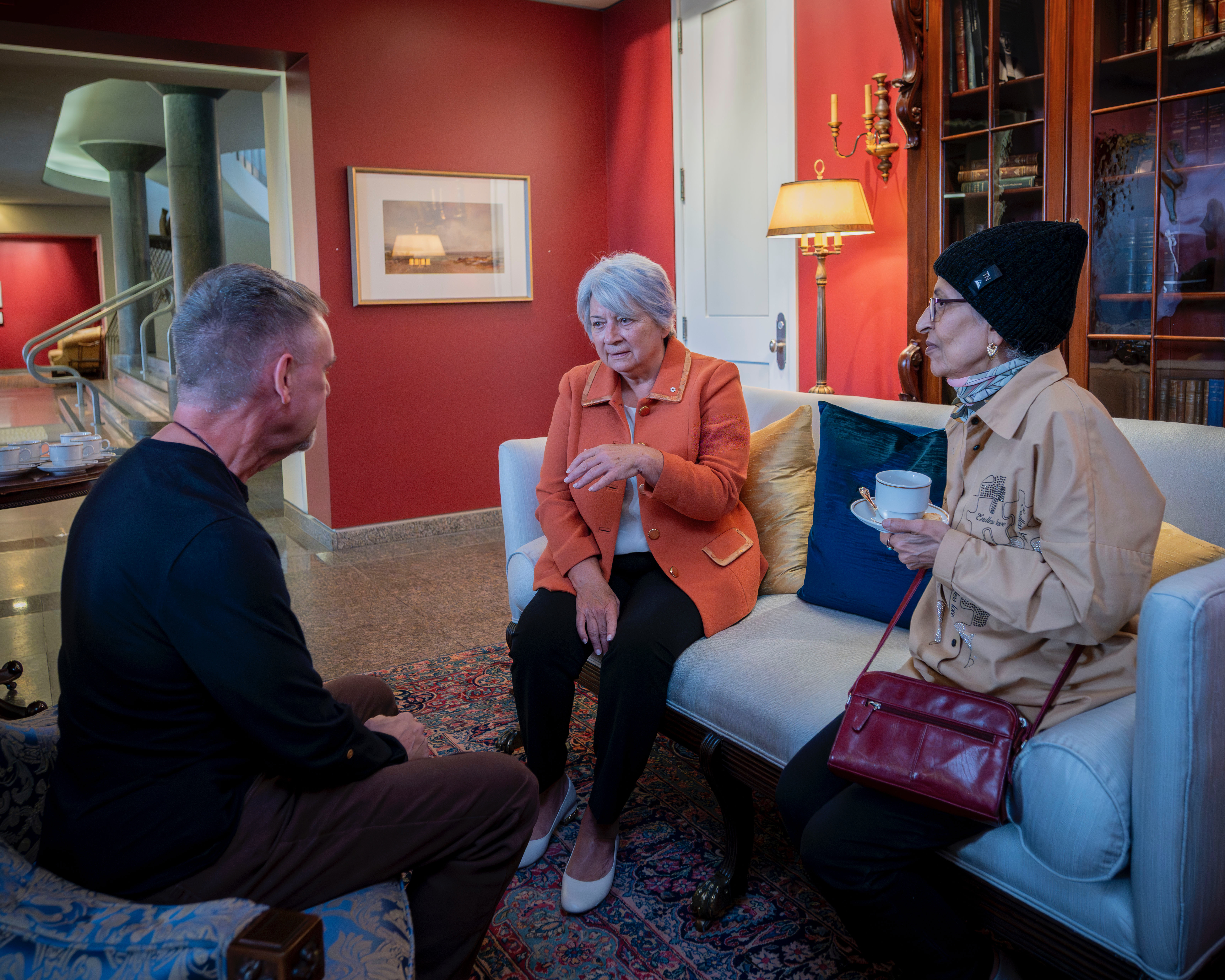 Un groupe de personnes est réuni dans une pièce chaleureusement éclairée, assis sur des canapés à motifs. La gouverneure générale Mary Simon est en pleine conversation. Quelques personnes se tiennent debout à l'arrière-plan, près d'une table où sont dispo