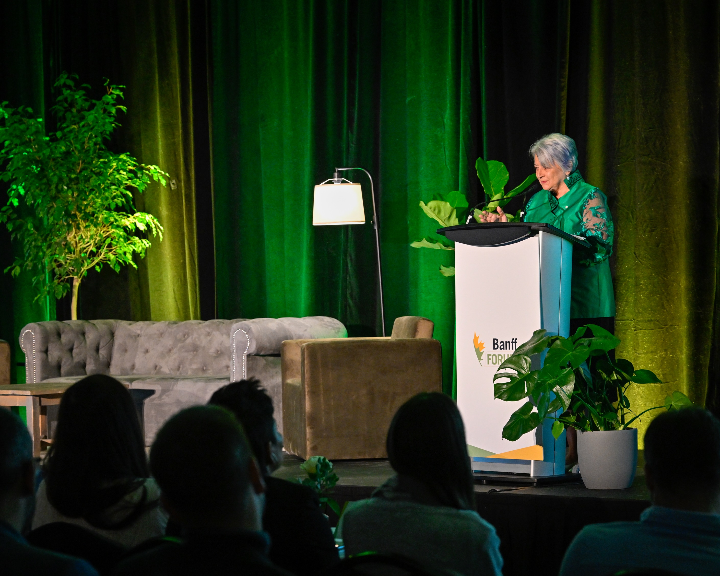 La gouverneure générale Mary Simon s'exprimant à un pupitre portant le logo « Banff Forum ». Elle se trouve sur une scène avec des rideaux verts, un canapé, des plantes et une lampe.