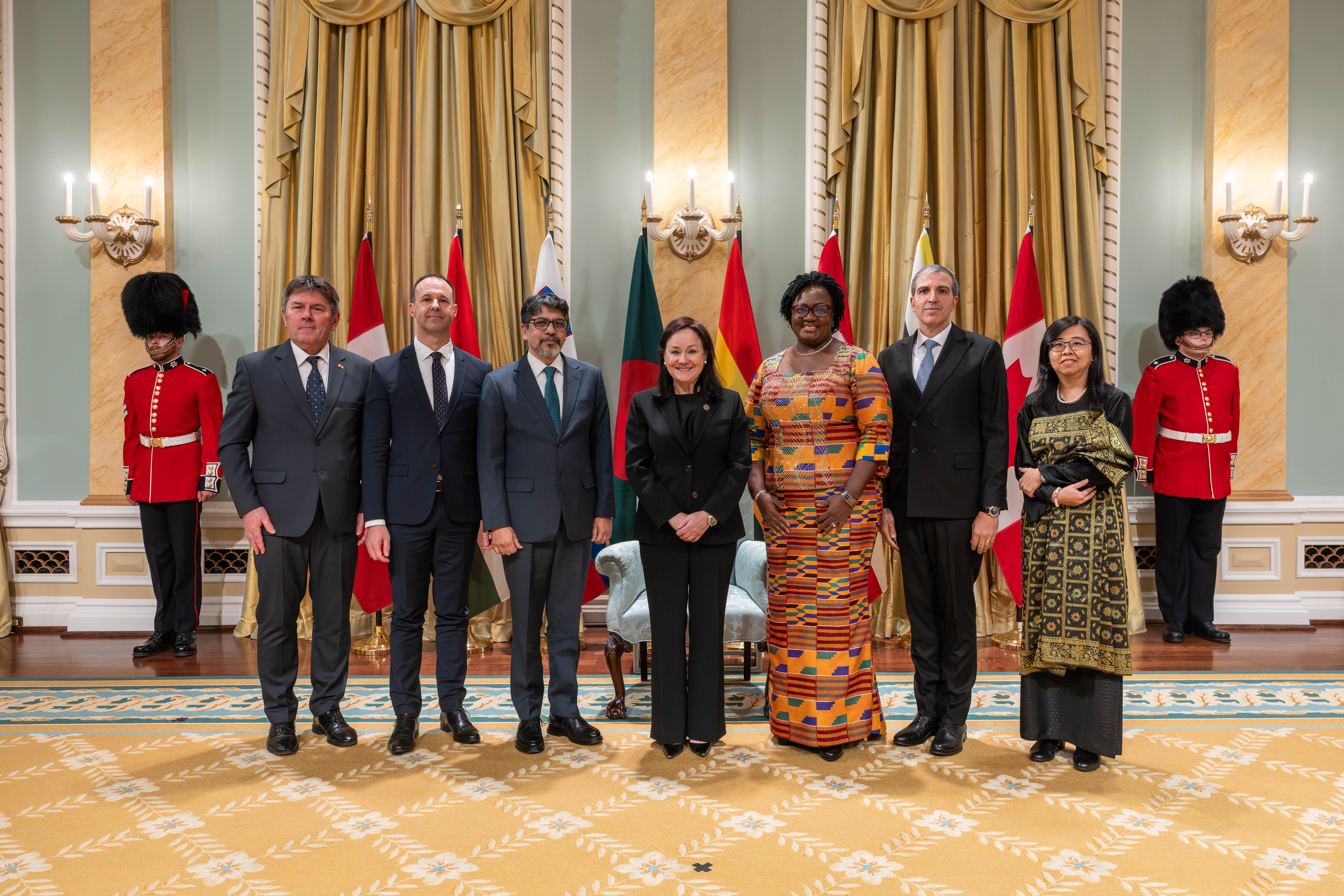 Photo de groupe avec l'honorable Suzanne Côté et six nouveaux chefs de mission, avec les drapeaux de chaque pays derrière eux. Deux gardes d'honneur se trouvent à l'arrière.