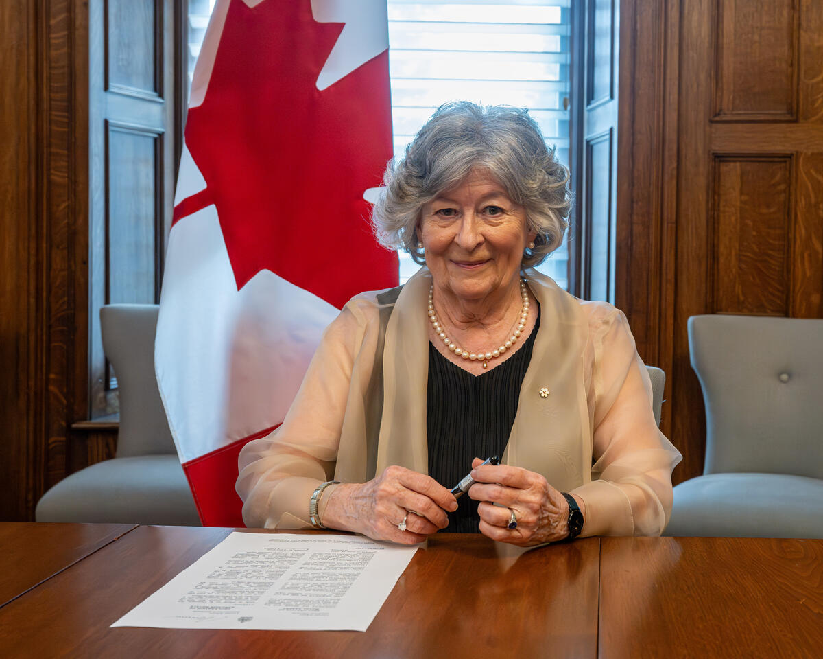 La gouverneure générale Arbour tient un stylo. Une feuille de papier se trouve devant elle, posée sur une grande table en bois. Un grand drapeau canadien est accroché derrière elle.