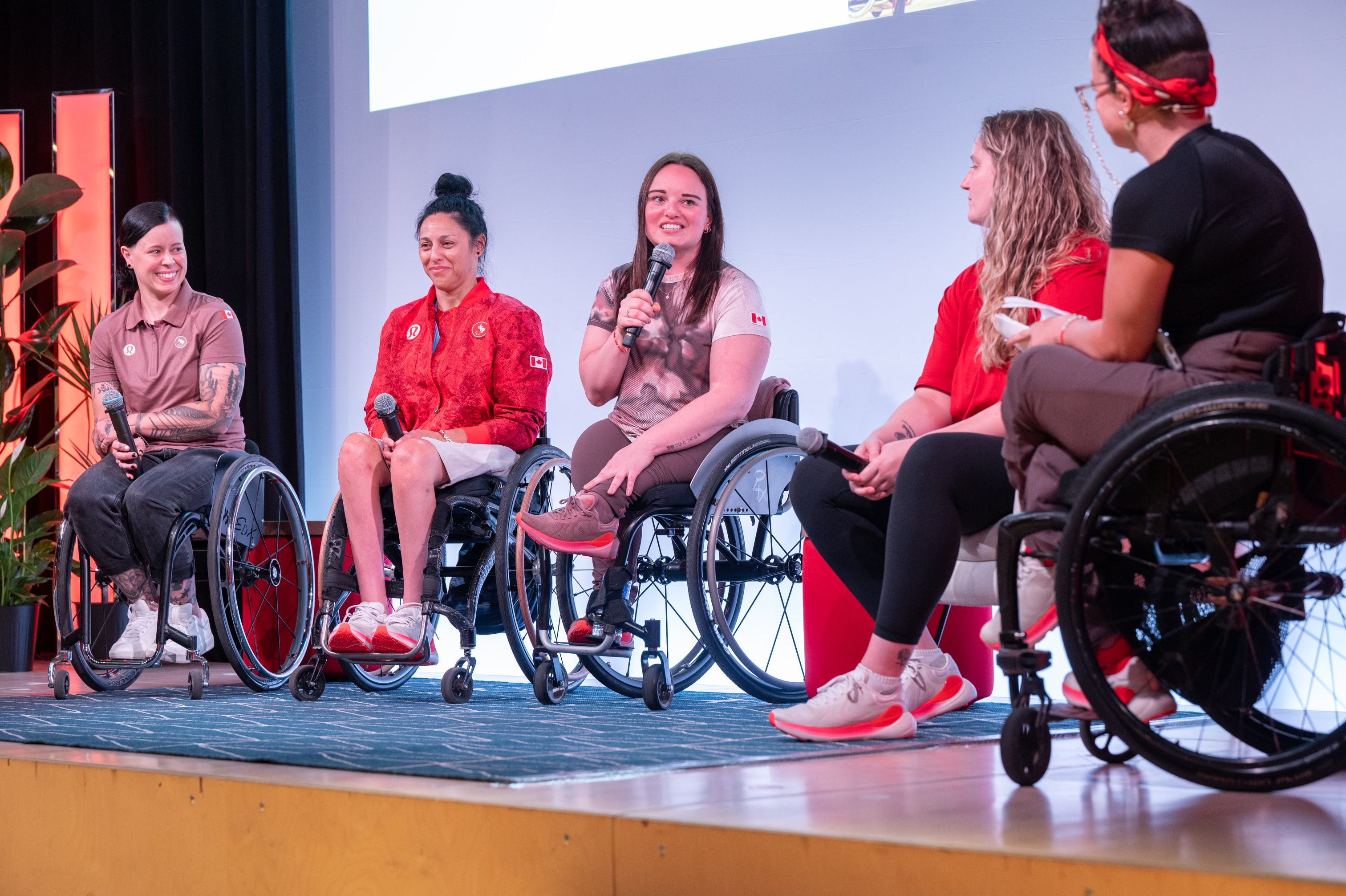Cinq personnes sont sur une scène, dont quatre en fauteuils roulants. Elles sont engagées dans une conversation ; une personne tient un microphone et parle, tandis que les autres écoutent et sourient.