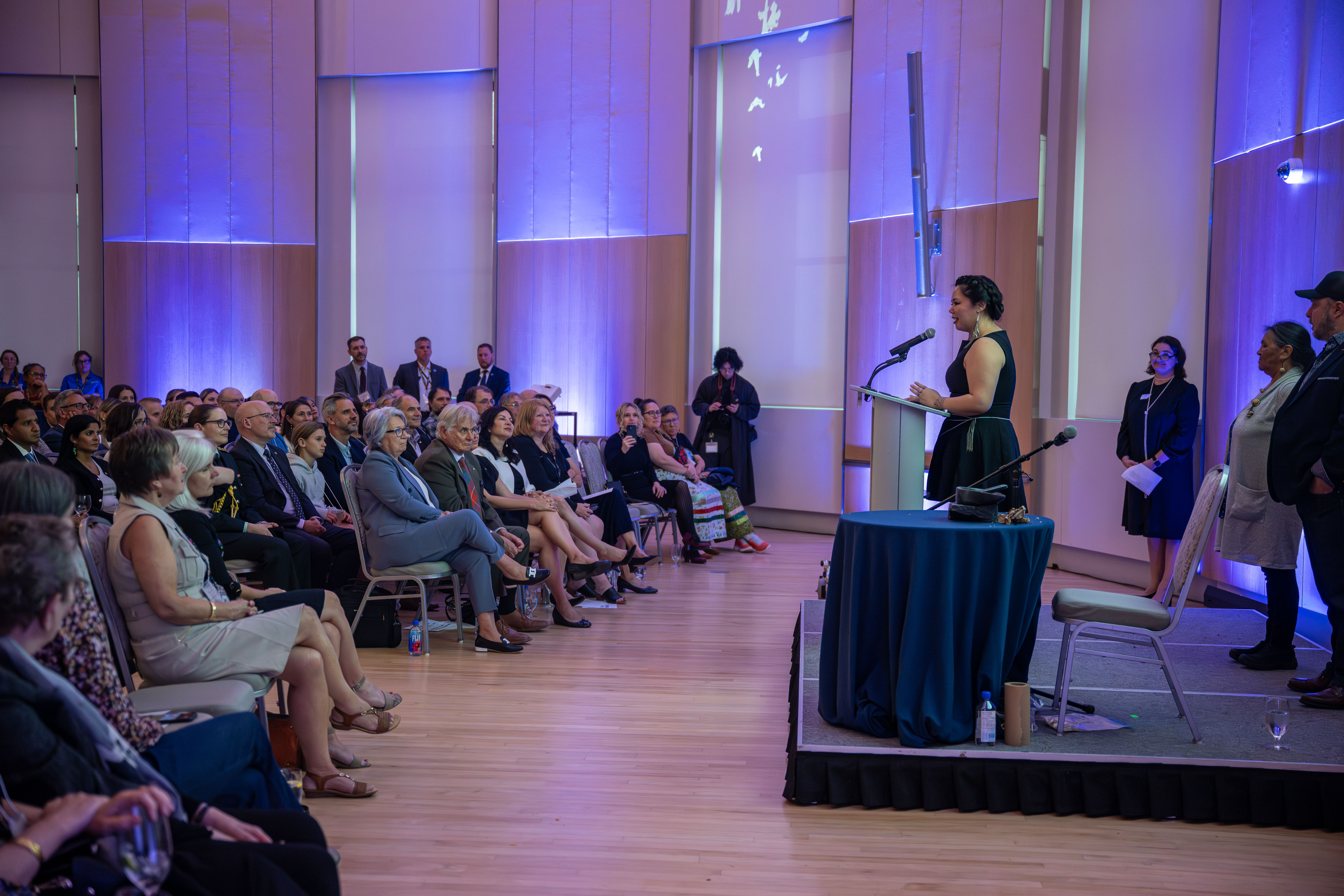A person standing on a small platform speaking to a seated audience.
