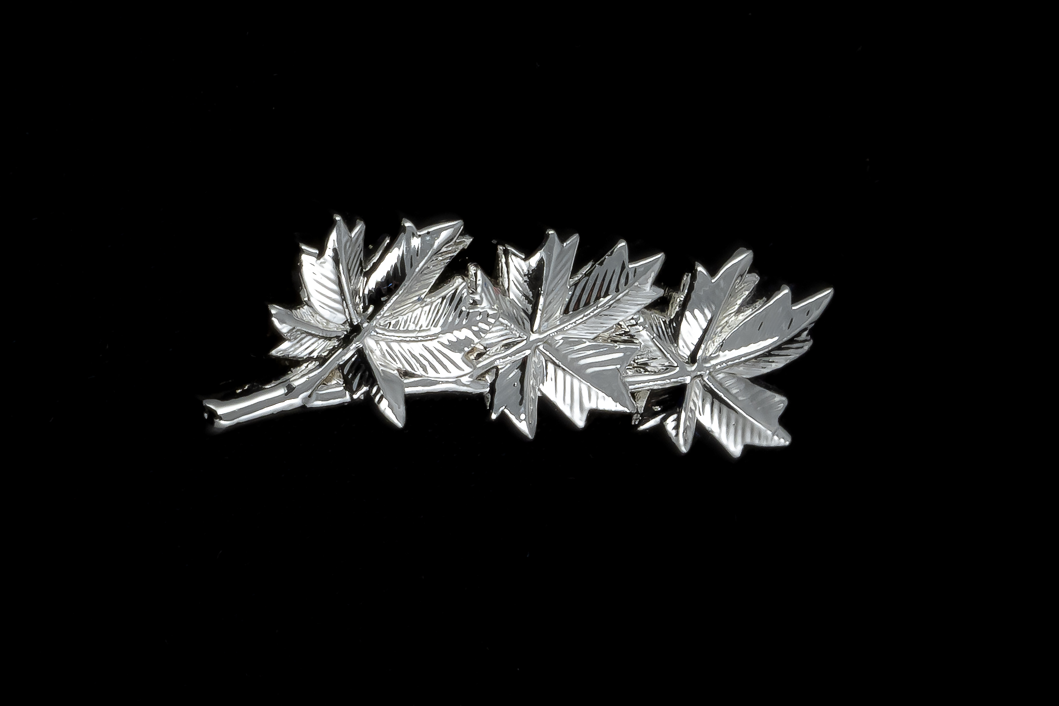 A silver pin on a black background. The pin consists of 3 maple leaves laying on top of the other.
