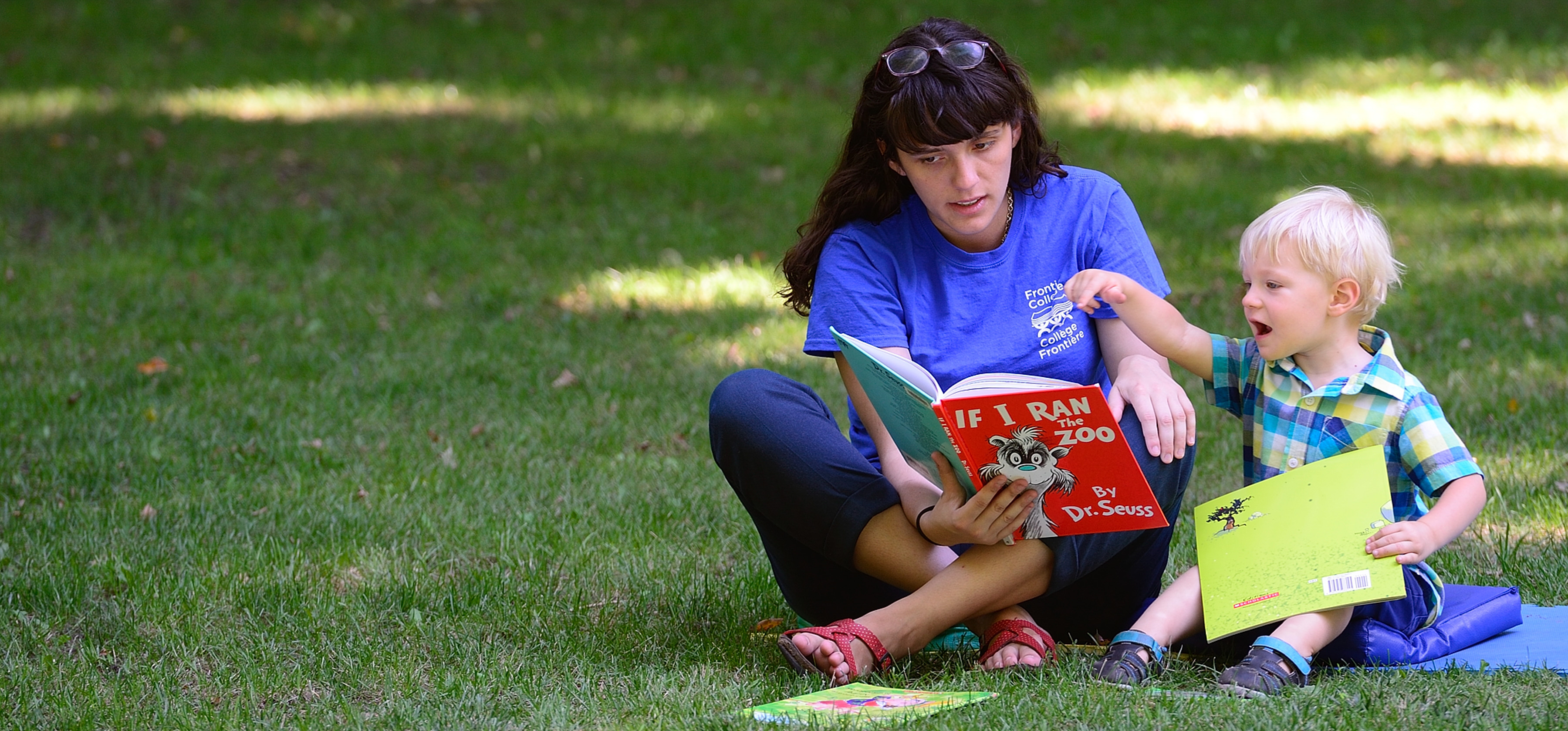 A woman reading a book to a little boy. They are seated on a blue blanket on the grass outside.