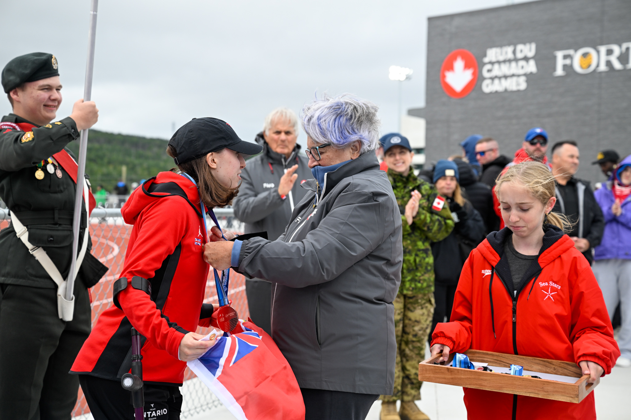 La gouverneure générale Mary Simon passe une médaille autour du cou d'une autre femme. Une jeune fille tient un plateau sur lequel sont posées des médailles.