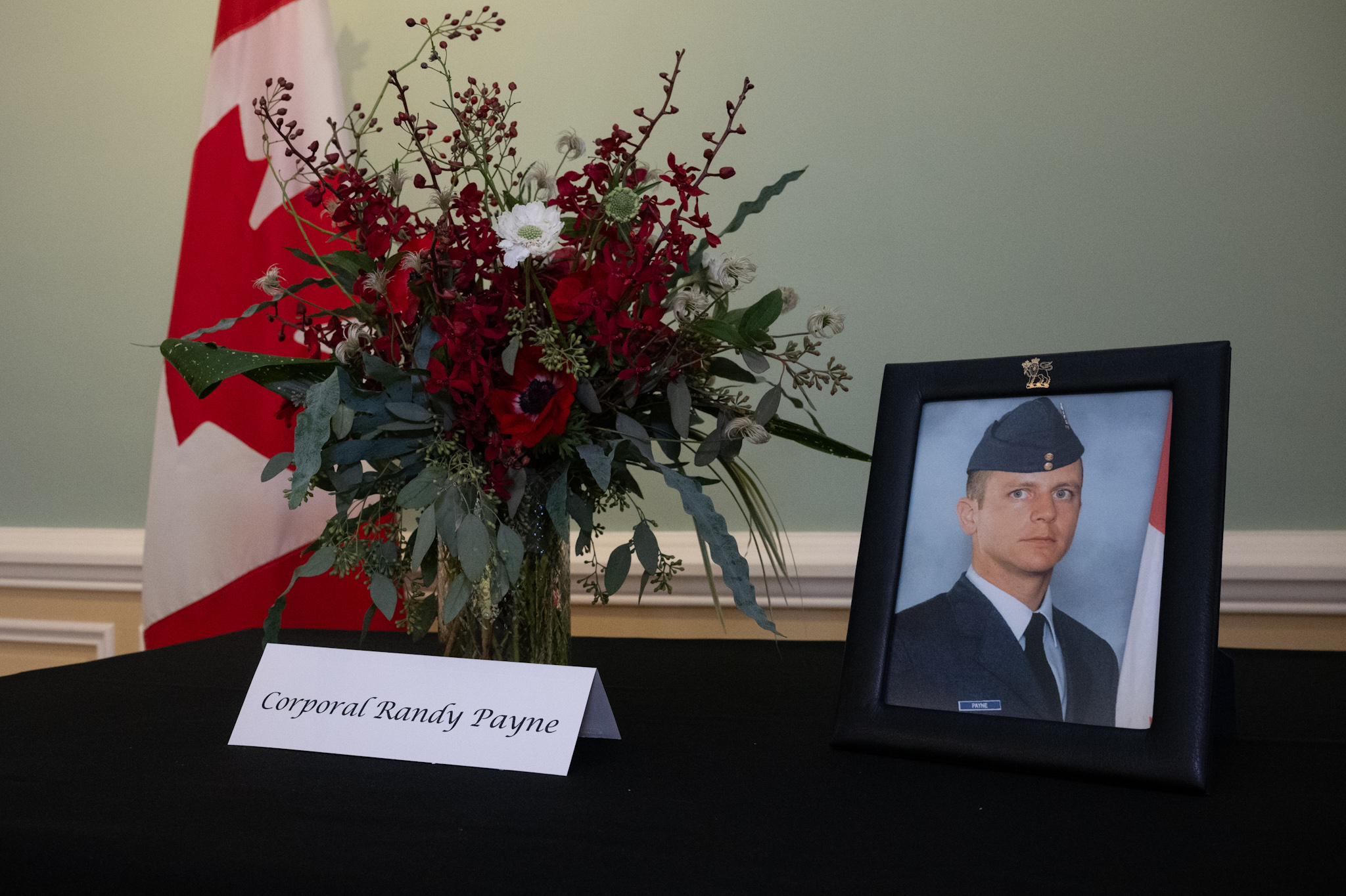 A framed photo of a military member on a table. A bouquet of flowers is beside the photo. A Canada flag is in the background. The name 'Corporal Randy Payne' is on a folded note card.