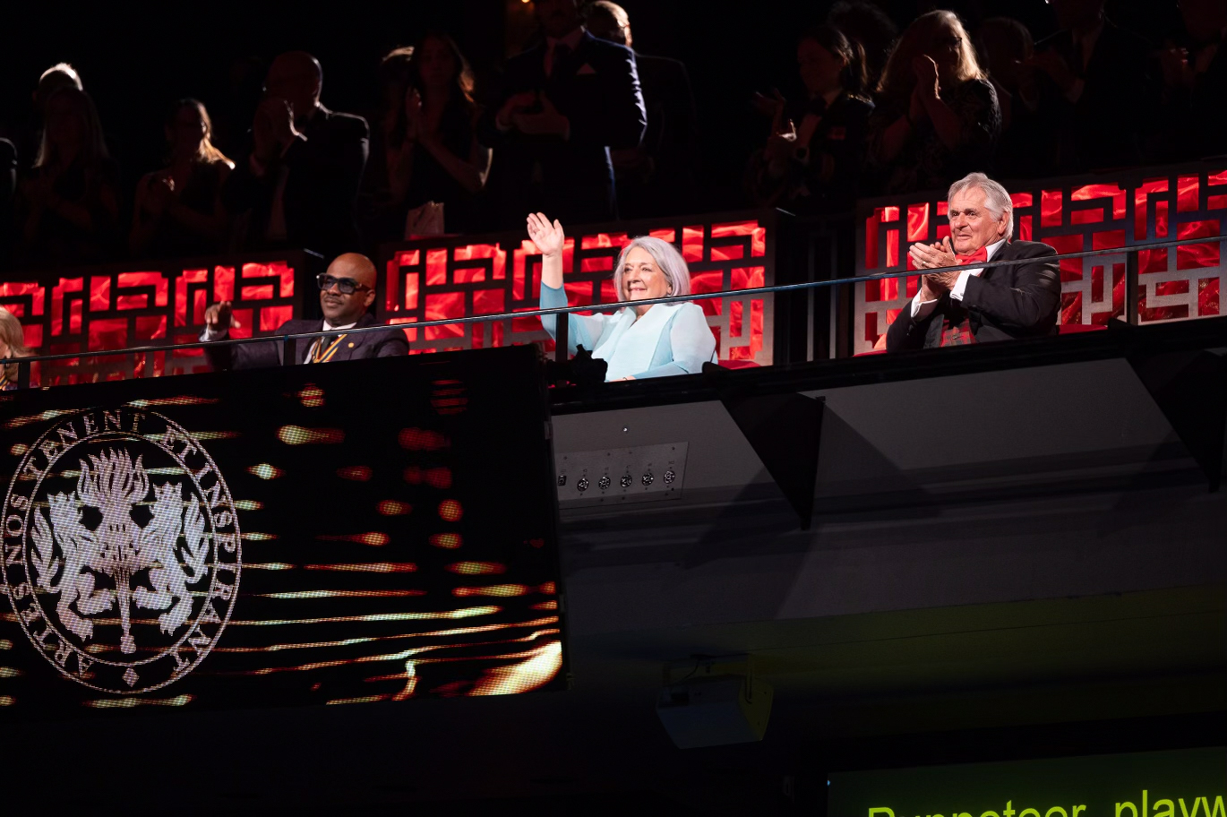 Governor General Mary Simon waves to the audience, seated next to Mr Whit Fraser and Wes “Maestro” Williams. The gala logo can be seen on a screen in front of them.