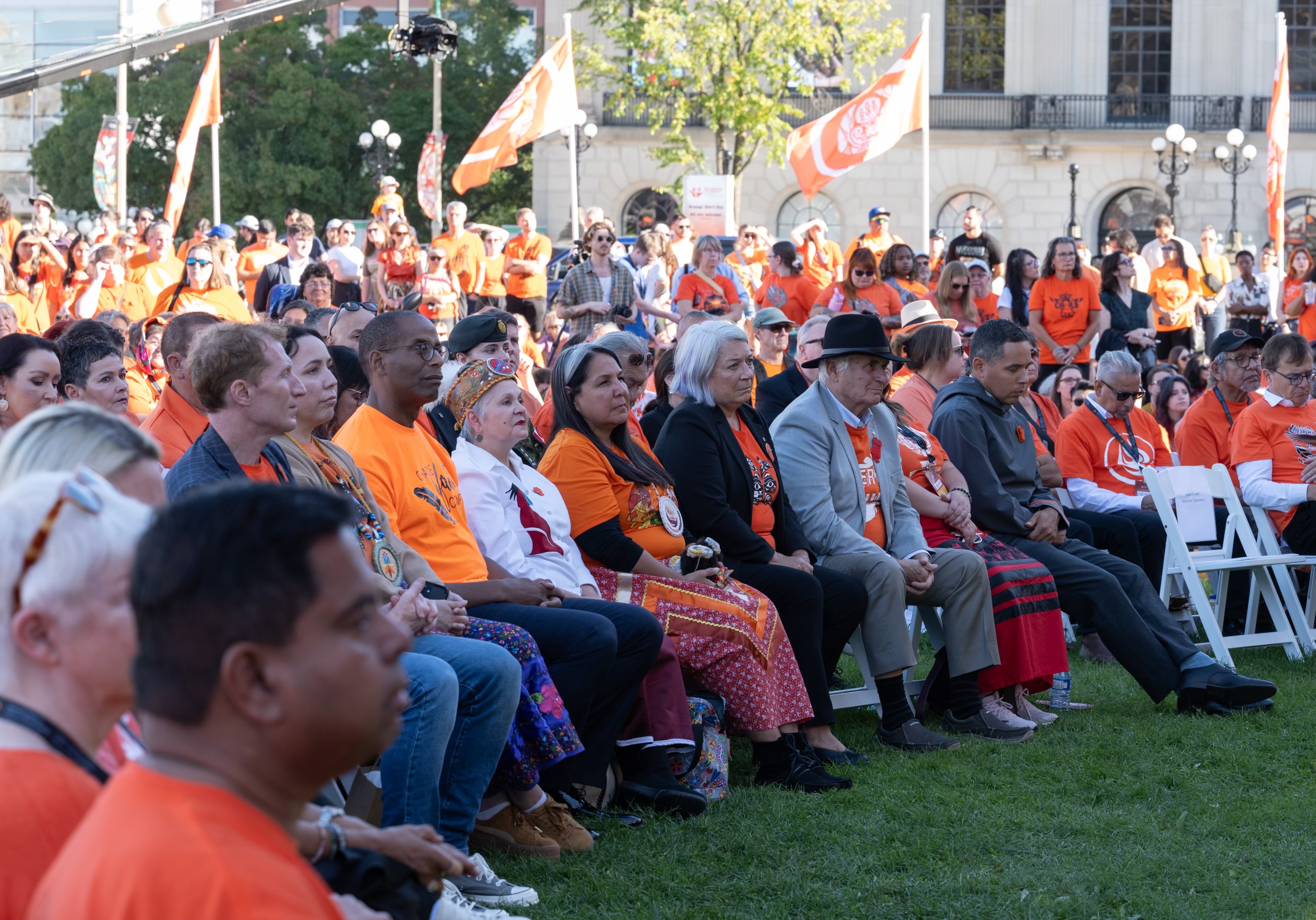 La gouverneure générale Simon et M. Fraser sont assis avec une grande foule de personnes sur des chaises à l'extérieur. Beaucoup portent des chemises orange et il y a des drapeaux orange à l'arrière-plan.