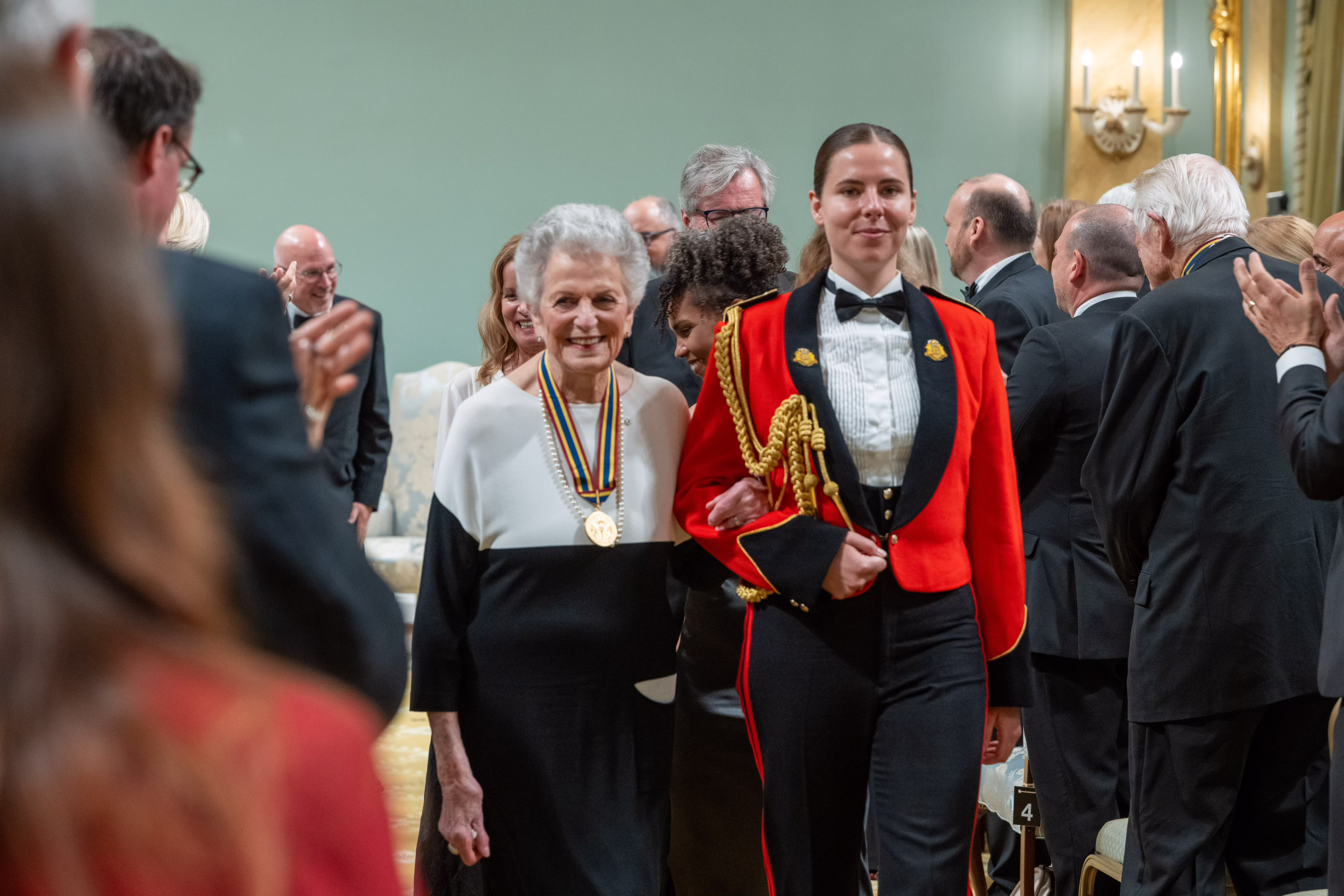Jenny Belzberg, accompanied by a member of the Canadian Armed Forces in a red uniform, walks out of the Ballroom at Rideau Hall through the audience