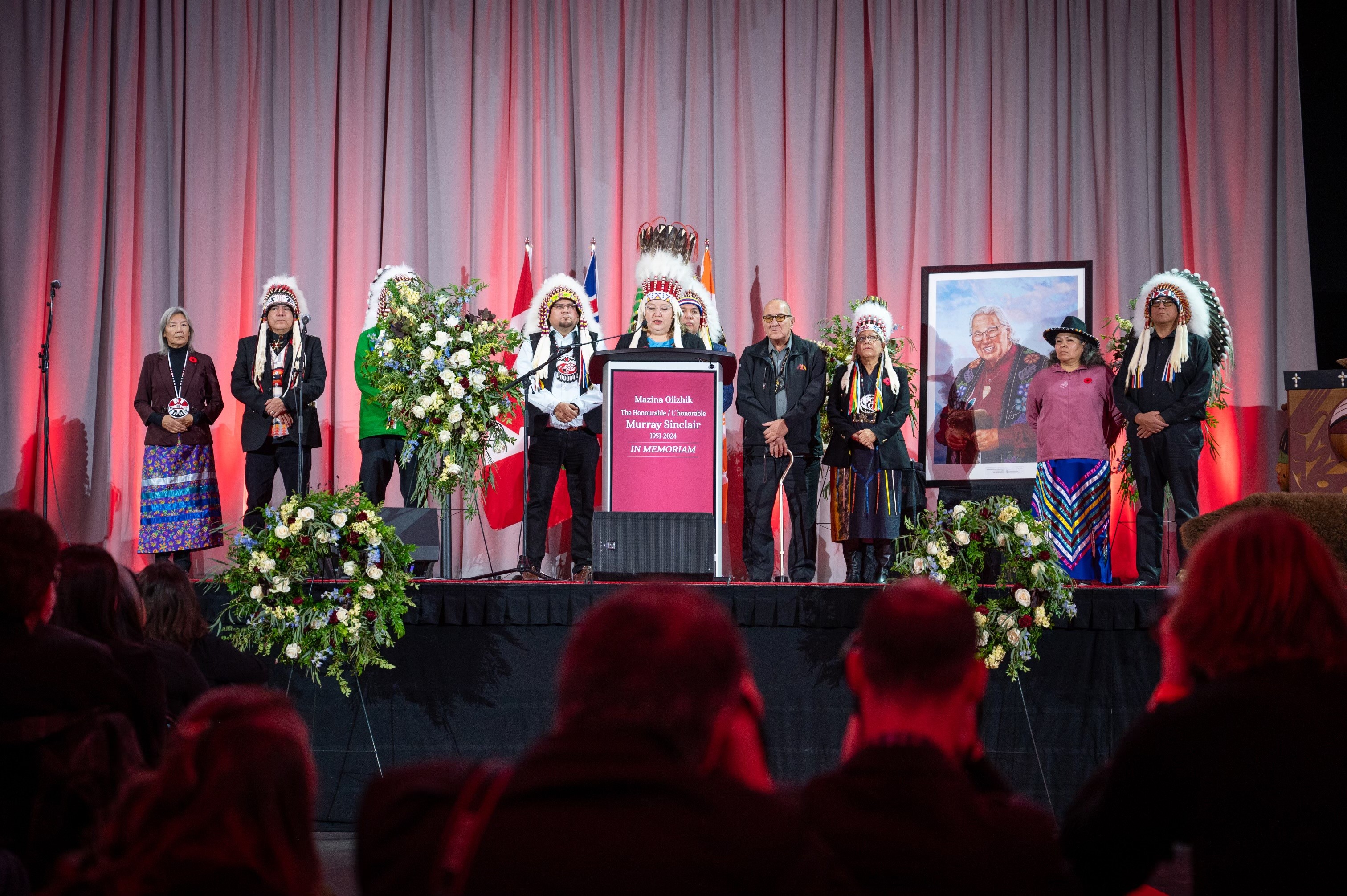 A group of Indigenous peoples, some of whom are wearing traditional clothing, are on a stage. One is speaking at a podium. We see the painted portrait of Murray Sinclair behind them.