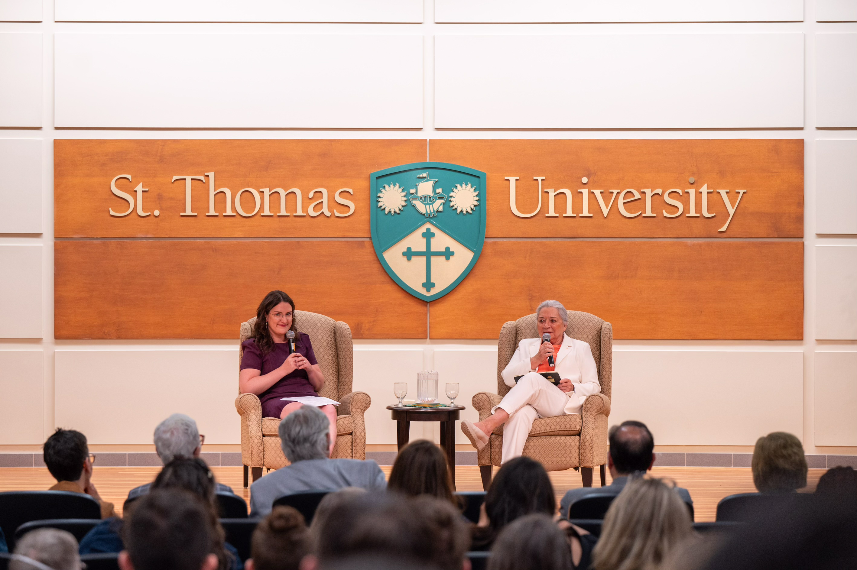 La gouverneure générale Simon et une autre personne sont assises sur des chaises longues sur la scène. Tous deux ont un micro. Les mots « St. Thomas University » sont sur le mur derrière eux.