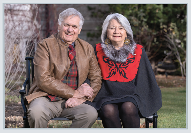 Mr. Whit Fraser and Governor General Mary Simon are sitting on a park bench, outside, smiling and holding hands, on a sunny fall day. M. Whit Fraser et la gouverneure générale Mary Simon sont assis sur un banc de parc, à l’extérieur. Ils se sourient et se tiennent la main. C’est une journée d’automne ensoleillée.