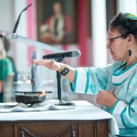 An Inuk Elder wearing traditional clothing holds her hand above a lit qulliq (oil lamp) as part of its inaugural lighting ceremony at Rideau Hall.