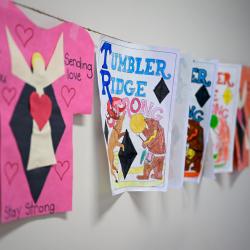 Un mélange de t-shirts découpés dans du papier, sur lesquels figurent des mots d'encouragement, et de simples morceaux de papier portant l'inscription « Tumbler Ridge Strong », accrochés les uns à côté des autres sur un mur.