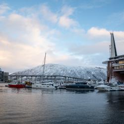 Plusieurs bateaux et yachts sont amarrés dans un port avec des bâtiments modernes et une montagne enneigée en arrière-plan, sous un ciel partiellement nuageux.