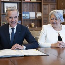 Le premier ministre Mark Carney et la gouverneure générale Mary Simon sont assis à une grande table en bois. Mark Carney signe un document.