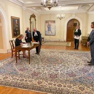 Vue de côté. Assise sur une chaise de bois, à une table de bois, la gouverneure générale signe un document.