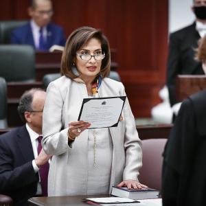 A woman dressed in white reads out loud from a document in her hands. She is reading to a woman wearing judge's robes who is standing across a medium-sized table from her.