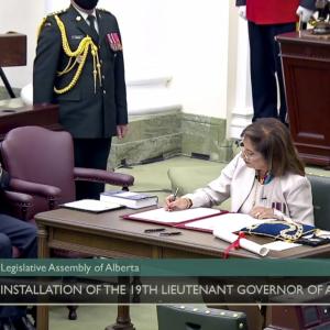 A woman dressed in white sits at a table. She is signing a document. Looking on are a man in a dark suit and a military member in service dress.