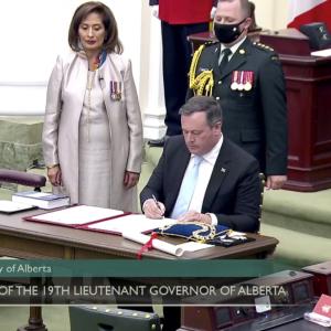 A man in a dark suit sits at a table. He is signing a document. Looking on are another man in a dark suit, a woman dressed in white and a military member in service dress.