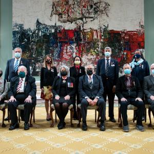 A group of eleven people, six sitting at the front and five standing behind, are posing for a photo in front of a large painting.
