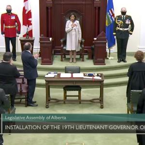 A woman dressed in white stands in front of a wooden throne flanked by three military members. She is facing a room full of applauding audience members who are facing her.