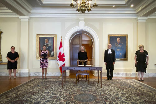 Cinq personnes sont alignées contre un mur jaune pâle. Une table est devant la personne au centre. Un drapeau du Canada se trouve à sa droite.
