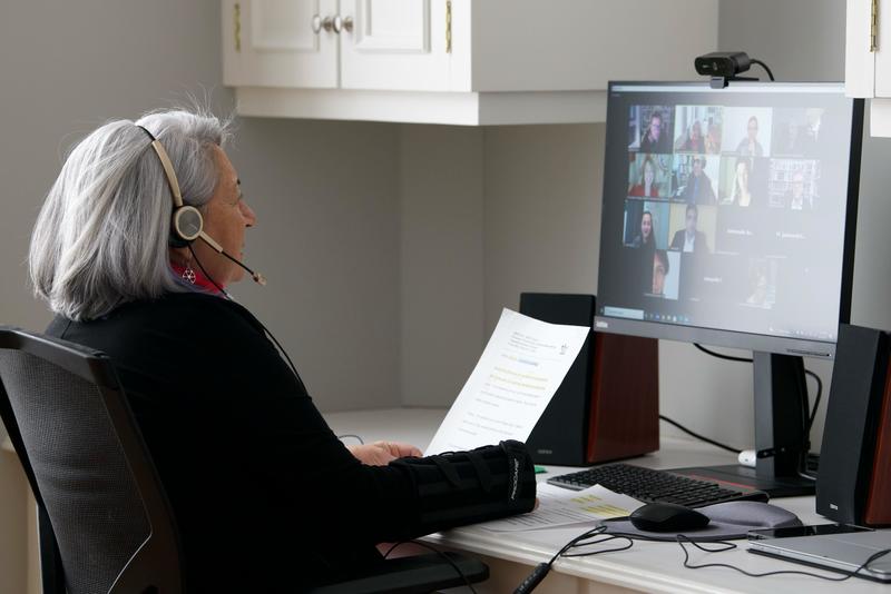 La gouverneure générale est assise à son bureau. Elle porte un casque d'écoute et participe à un événement virtuel sur son ordinateur. Elle tient une feuille de papier.