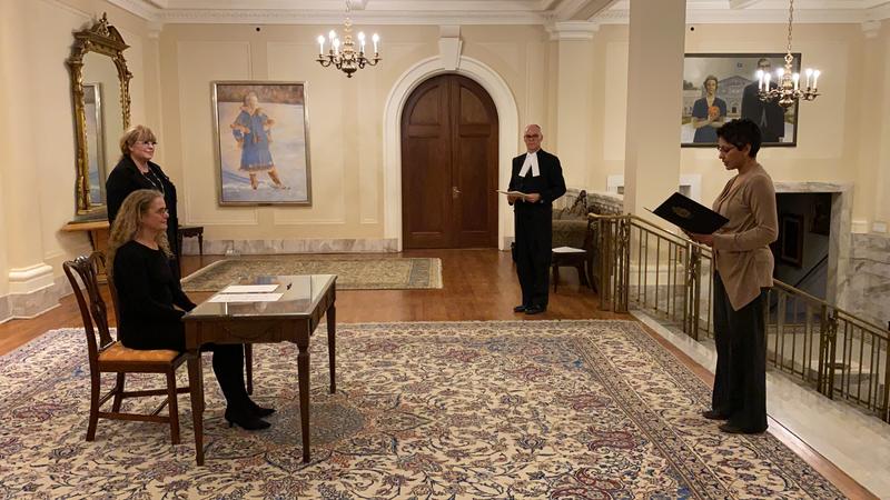 The Governor General is sitting at a desk. On the right hand side, there is a man holding a document in his hands. Four other people are standing around the desk all while respecting the social distancing norms. 