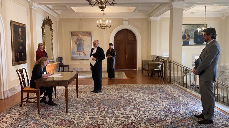 The Governor General is sitting at a desk. On the right hand side, there is a man holding a document in his hands. Four other people are standing around the desk all while respecting the social distancing norms. 