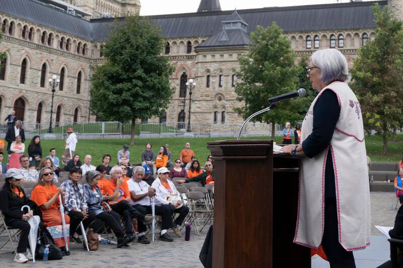 La gouverneure générale Mary Simon est debout derrière un pupitre. Des gens sont assis devant elle. Plusieurs personnes portent des chandails orange. Un grand bâtiment en pierre se trouve à l'arrière-plan.
