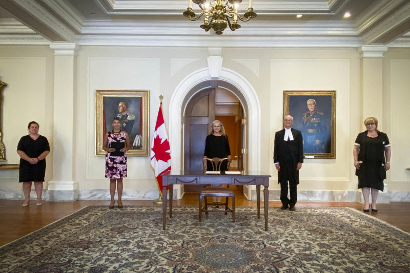 Five people standing in front of a pastel yellow wall. A table is in front of the person standing in the middle. A Canadian flag is placed to her right.
