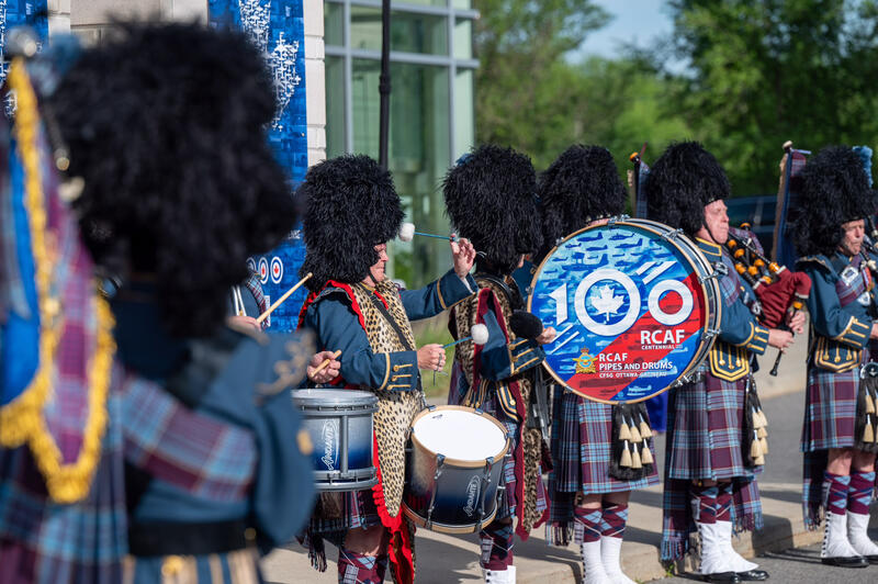 Des joueurs de tambour et de cornemuse en uniforme militaire se produisent à l'extérieur. Sur un grand tambour, on peut lire, en anglais, «100 RCAF» (100 ARC).