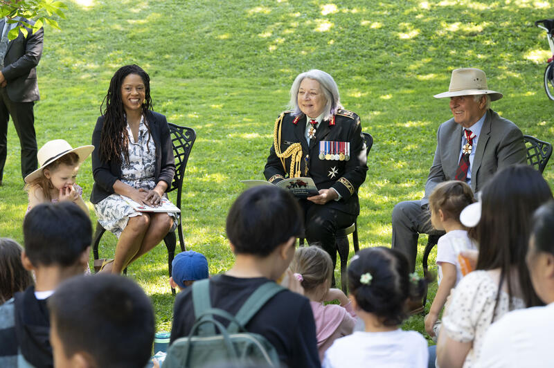 La gouverneure générale Mary Simon sourit à la foule. Elle tient un livre et porte l'uniforme de l'Armée canadienne. Une femme est assise à sa droite et M. Whit Fraser est assis à sa gauche. Il y a des enfants au premier plan.