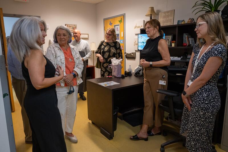  A group of five people converse in an office, surrounded by shelves and a desk.