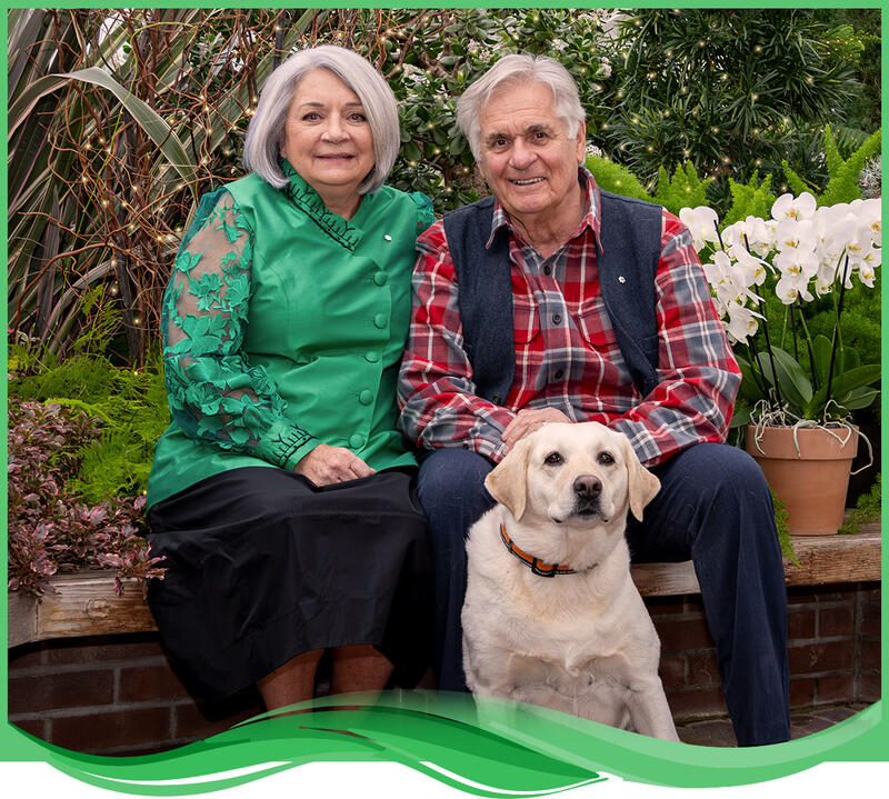 The Governor General and Mr. Fraser pose with their dog. They are in a greenhouse surrounded by a background of plants.