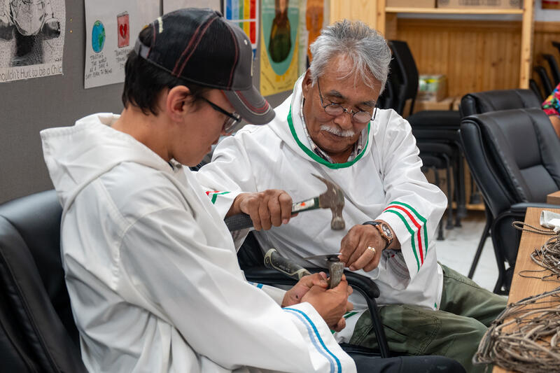Two people seated on a couch. One of them is using a hammer.