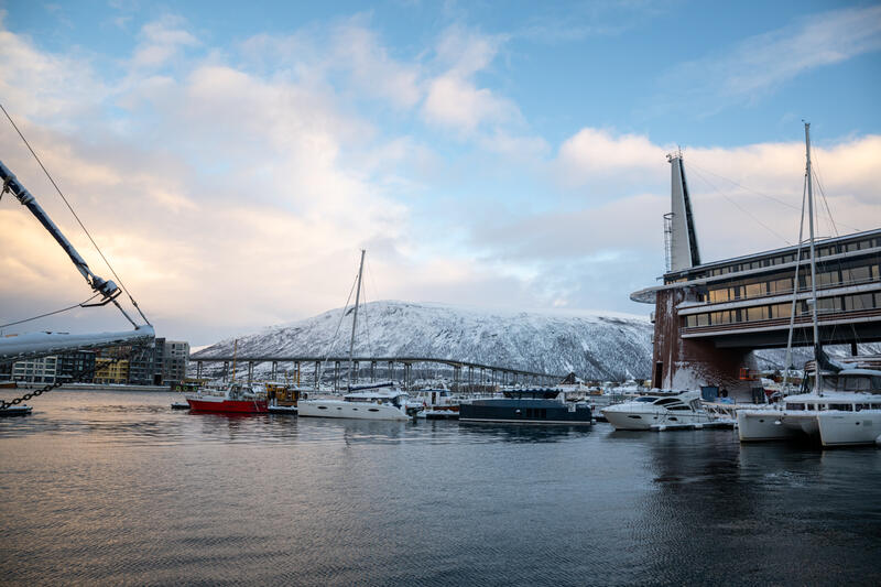 Plusieurs bateaux et yachts sont amarrés dans un port avec des bâtiments modernes et une montagne enneigée en arrière-plan, sous un ciel partiellement nuageux.