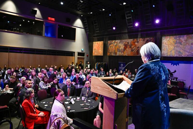 Governor General Mary Simon is seen from the back, standing, speaking at a podium, on a stage, in front of a room filled of people, sitting at round tables.