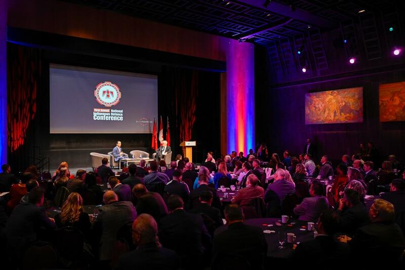 A large dark room, with a lighted staged on which two man are seated in chairs. On a screen behind them is written First annual National Indigenous Defence Conference under a red, orange and white logo with a red bird in a circle.