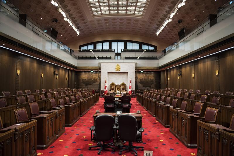 Empty Senate of Canada chamber. Several dark desks down the middle aisle. Dark brown leather chairs in seating areas on either side. A large throne at the front of the room. A glass-enclosed balcony overlooks the chamber.