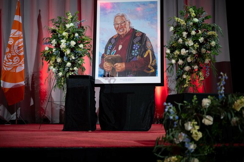 Painted portrait of Murray Sinclair showcased on a stage covered with red carpet ,surrounded by funeral flowers.arrangements.