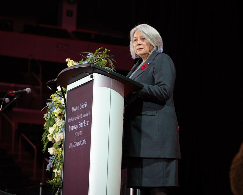 Governor General Mary Simon is speaking at a podium. She wears a grey suit, with a poppy. A funeral flower arrangement is on the left of the podium.