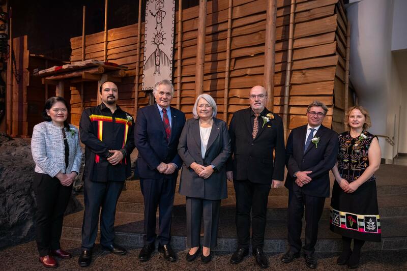 Governor General Mary Simon and Mr. Whit Fraser are standing in the centre of a line of 5 other people, in front of a wooden wall. Everyone is smiling at the camera.