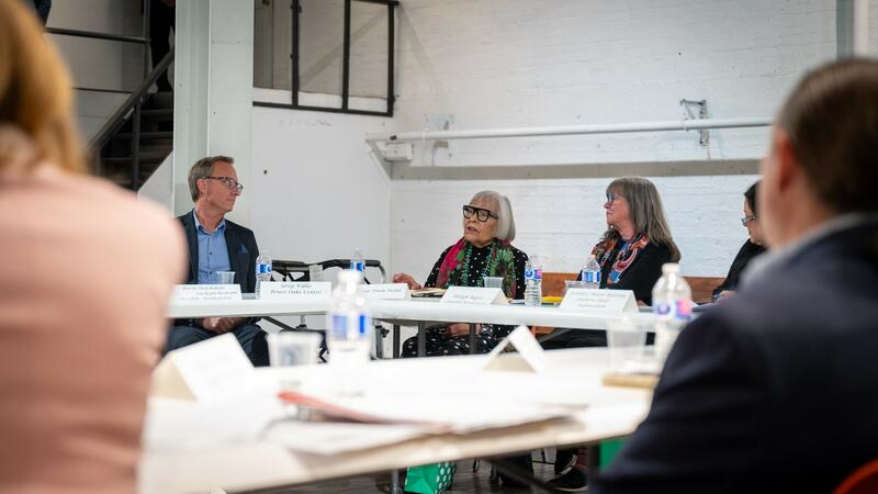 A group of people sit at a conference table in discussion. Papers and water bottles are on the table.